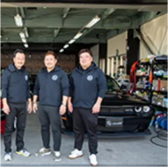 ガレージの前で黒いフード付きパーカーを着た3人の男性が車と共に並んで立っている写真。