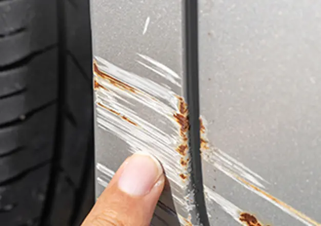 自動車のボディにできた傷と錆を指差している様子、塗装の剥がれが確認できる