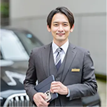 スーツ姿の男性がタブレットを手に微笑みながら立っている、背景には高級車