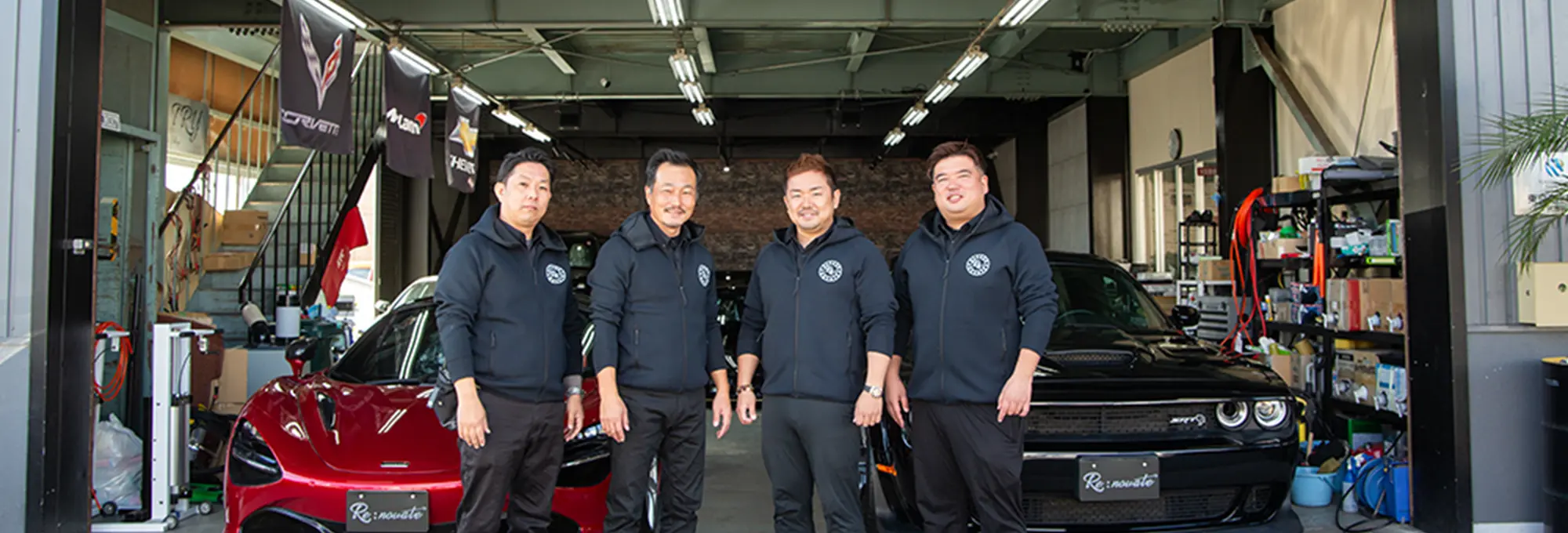 自動車整備工場の前で、黒いパーカーを着た4人のスタッフが並んでポーズを取っている。